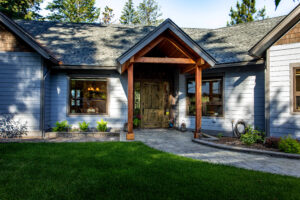 A modern house with blue-gray siding, a wooden front door, and a peaked porch supported by wooden beams—perfect for those seeking Bigfork Real Estate. Enjoy a green lawn, landscaped garden beds, and tall trees in the background.