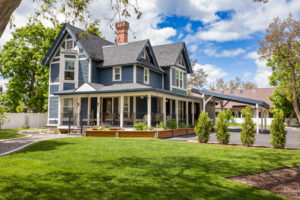 A large, two-story blue house with white trim and a wraparound porch sits on a manicured lawn with trees and shrubs—an inviting example of Kalispell Real Estate under a bright, partly cloudy sky.