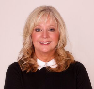 A smiling woman with wavy blond hair, wearing a black top with a white collar, photographed against a plain light background—your trusted Kalispell Realtor.