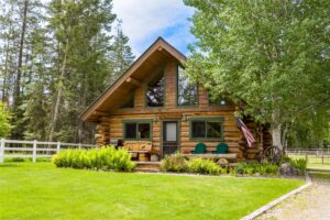 A cozy log cabin with large windows and a front porch sits among green grass and trees—perfect for enjoying Flathead Valley Real Estate. Two chairs, a bench, and an American flag complete the charm, with a white fence lining the background.