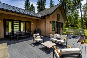 A modern wooden house with a spacious outdoor deck featuring cushioned chairs, tables, and a dining area—perfect for relaxing in nature. Surrounded by tall trees, it's an inviting retreat often showcased by Bigfork Real Estate professionals.