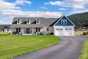 A modern two-story house with white siding and blue accents, featuring a front porch, dormer windows, and a double garage. The manicured lawn and curved driveway reflect intention and new beginnings amid hills and a blue sky backdrop.