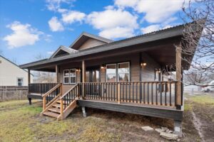 A modern dark wood house with a large covered front porch, wooden railings, and steps, set on a grassy yard bursting with spring colors under a blue sky with scattered clouds.