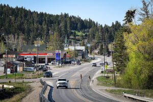 A two-lane road curves through a small town with shops, cars, and a gas station on the left. Trees and green hills surround the area, perfect for July Journeys. Two deer cross the road in the foreground.