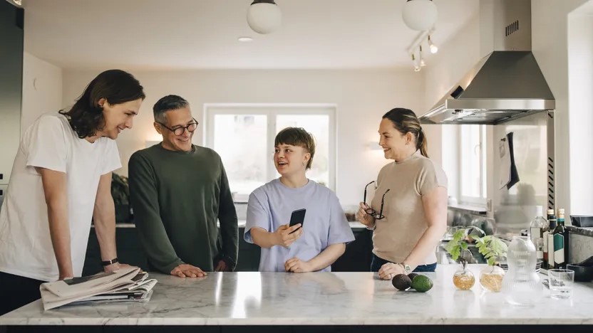 Four people stand around a kitchen island, smiling and chatting. One person holds up a smartphone, sharing something with the others. Various kitchen items and produce hint at cozy season gatherings in this bright, modern space filled with community spirit.