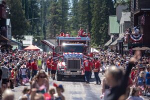 A patriotic parade with a large decorated truck drives down a street lined with cheering crowds, many dressed in red, white, and blue—celebrating both freedom and the hot real estate summer listing season beneath festive banners.