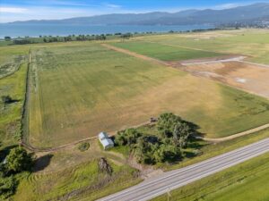 Aerial view of a large farmland with green and tan fields, a small white building, clusters of trees, a road in the foreground, and a distant lake with mountains—perfect for a winter real estate update under the bright blue sky.