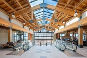 Spacious, modern interior with wooden beams, glass ceiling, and large windows. Two escalators descend to a lower level, with seating areas on both sides and natural light filling the space.