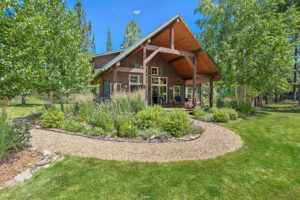 A cozy wooden cabin with a covered porch sits amid lush green grass and landscaped gardens under a bright blue sky—a true Flathead Valley Real Estate gem. Gravel pathways curve through the garden in front of the house.