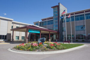 A modern hospital building with a main entrance canopy, three flagpoles displaying flags, and a circular flower bed with colorful tulips in the foreground under a clear blue sky.