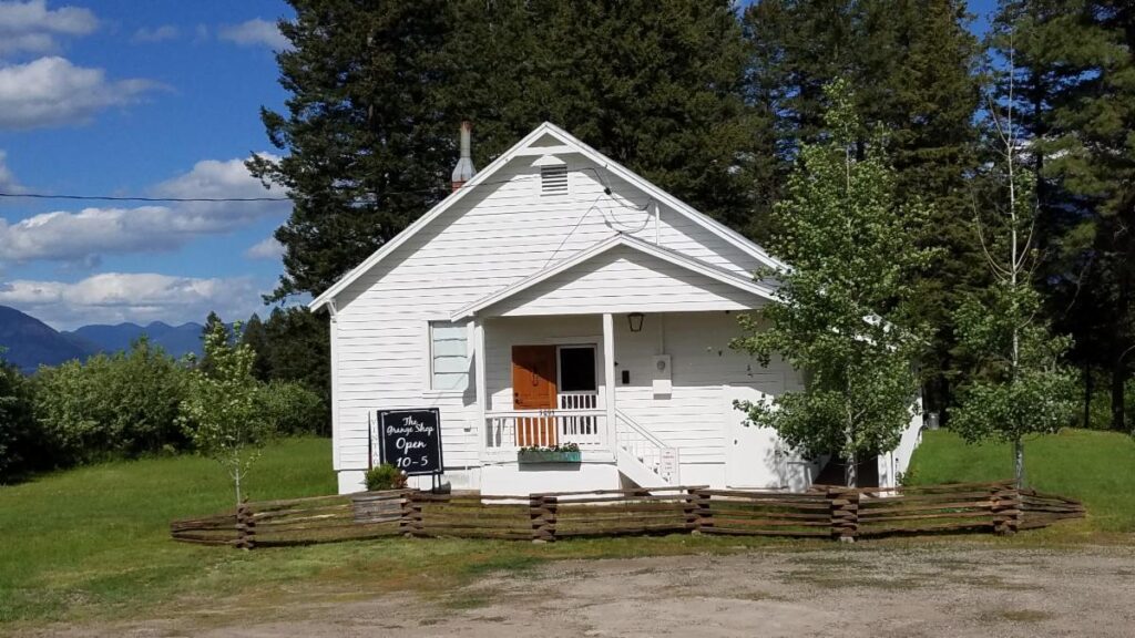 A small white building with a porch and open door sits behind a rustic wooden fence, surrounded by full green grass and rolling trees under an August blue sky. A sign near the entrance reads, Open 10-5.
