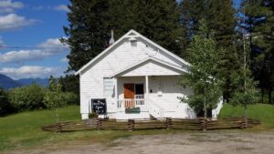 A small white building with a porch and open door sits behind a rustic wooden fence, surrounded by full green grass and rolling trees under an August blue sky. A sign near the entrance reads, Open 10-5.