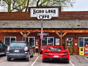 A rustic wooden building with a sign reading Echo Lake Cafe sits under soft February Light. Two cars, including a black SUV and a red sports car, are parked in front. An American flag hangs beside the entrance, and the cafe displays an OPEN sign.