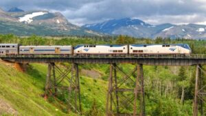 A passenger train crosses a tall steel bridge over a green valley, with lush trees and mountains capped with patches of snow in the background—an enchanting scene steeped in local folklore beneath a cloudy sky.