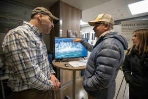 Two men stand by a table with a monitor displaying a 3D model of a building; one man points at the screen while the other listens. A woman stands nearby, observing the conversation as they share ideas for a Bright New Year.