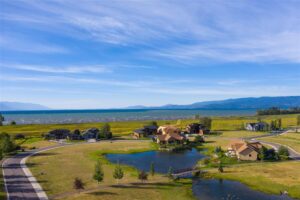 Aerial view of houses near a small pond, with a large grassy field leading to a lake and distant mountains under a blue sky. A paved road curves through the peaceful neighborhood, where love and market energy are both in the air.