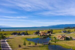 A cluster of houses sits near a small pond on grassy land, with a lake and distant mountains under a blue sky. A winding road suggests movement and new beginnings through the scenic, peaceful landscape.