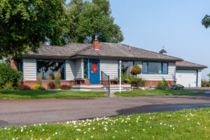 Single-story house with gray siding, red brick accents, large windows, and a bright blue front door. A well-kept lawn and small trees enhance the sense of new beginnings, while a paved driveway adds to the welcoming movement of the home.