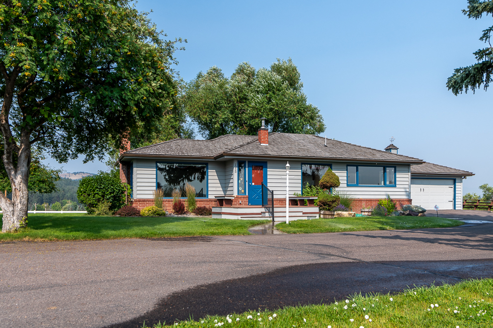 A single-story house with a gray exterior, blue front door, red brick accents, attached garage, and a well-kept yard graced with trees and shrubs in full Spring bloom on a sunny day.