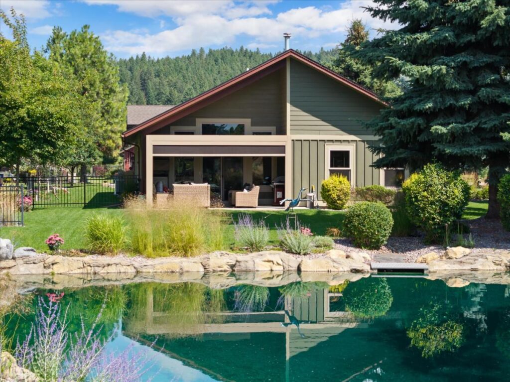 A green house with large windows faces a lush backyard, where a stone-edged pond reflects August Reflections among grass, shrubs, flowers, and tall trees under a blue sky dotted with clouds.