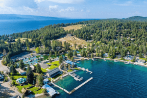 Aerial view of a lakeside community with docks, boats in movement, green trees, and houses surrounded by forested hills; blue lake water and distant mountains under a partly cloudy sky evoke new beginnings.