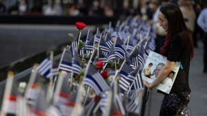 A woman stands by a memorial covered with small American flags and roses, holding a collage of photos and looking thoughtful amid a crowd, as the changing season brings cool crisp days of fall.