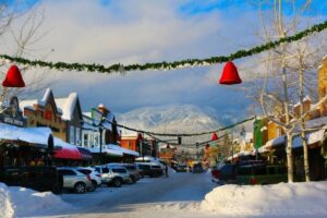 A snowy downtown street lined with shops and parked cars, decorated with green garlands and red bells, hints at new beginnings as movement flows beneath snow-covered mountains under a partly cloudy sky.