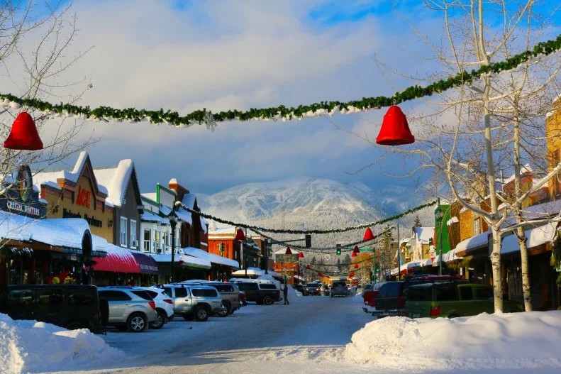 Snow-covered small town street with cars parked along both sides, festive garlands and red bell decorations hanging overhead for Bigfork Days, and mountains in the background under a partly cloudy sky.