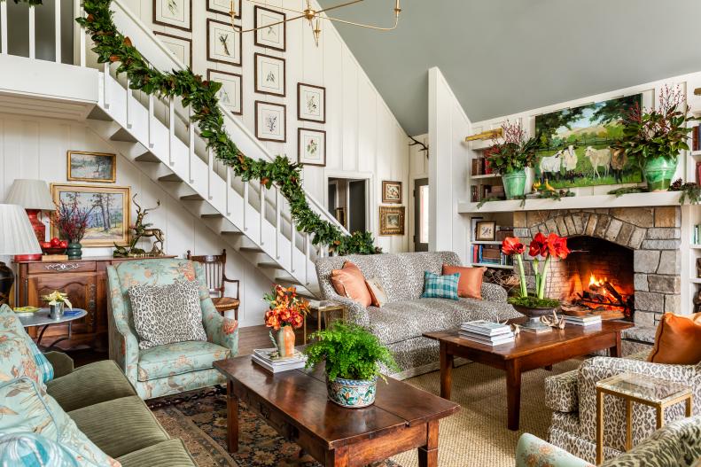 A cozy living room decorated for Christmas with garland along a staircase, a lit fireplace, patterned sofas, floral arrangements, and framed art on the walls. A festive, inviting atmosphere wishing you a Merry Christmas and a Bright New Year.
