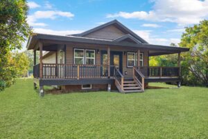 A modern single-story house with a large covered front porch, wooden railings, and steps welcomes you—perfect for enjoying the lush greenery and blue skies of Flathead Valley during the vibrant Spring Market season.