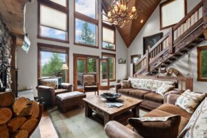 A cozy living room with large windows, rustic wooden furniture, a stone fireplace, log accents, and an antler chandelier sets the intention for new beginnings, offering views of trees and a wooden staircase leading upstairs.