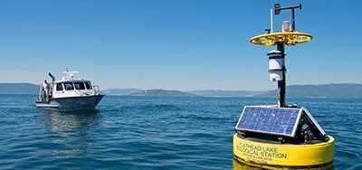 A yellow buoy with solar panels and scientific equipment floats on a calm lake in August, with a research boat nearby and rolling mountains visible in the background under a clear blue sky.