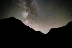 Silhouette of mountain peaks under a night sky filled with stars and the Milky Way galaxy, evoking cozy season vibes and a sense of community spirit amidst the dramatic contrast of the dark landscape and luminous night sky.