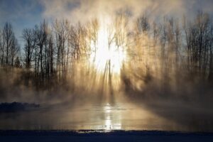 The sun shines brightly through bare winter trees, heating up the mist rising from a partially frozen river or lake, casting long shadows and creating a dramatic, ethereal scene.