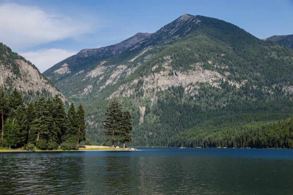 Embrace Lake Days by a calm lake surrounded by dense green pine forests, with tall, rugged mountains rising in the background beneath a blue sky with scattered clouds.