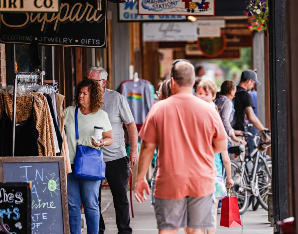 People walk along a busy sidewalk lined with shops, soaking in the cozy season vibes. Some browse clothing displays or carry shopping bags, while bicycles are parked nearby and a lively community spirit fills the air.