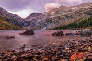 Rocky shoreline of a clear lake with large stones in the water, surrounded by pine trees and rugged, snow-dusted mountains under a pink and purple October sunset—a perfect scene for falling for fall.