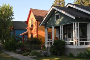 A row of colorful houses with porches and gardens, featuring green, yellow, and blue exteriors, basks in a winter glow under the evening sunlight with trees in the background—perfect for your December real estate update.