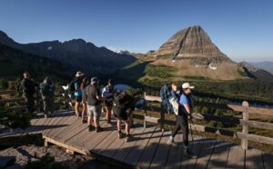 A group of hikers stands on a wooden overlook with backpacks, admiring a scenic mountain view under a clear blue sky. The community lights twinkle below, while a prominent, pyramid-shaped peak—steeped in local folklore—rises in the background.
