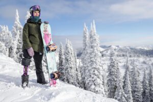 A snowboarder with a prosthetic leg stands on a snowy slope in Flathead Valley, holding a snowboard with a colorful design. Snow-covered trees and mountains form the backdrop under a partly cloudy sky, hinting at the excitement of spring market season.