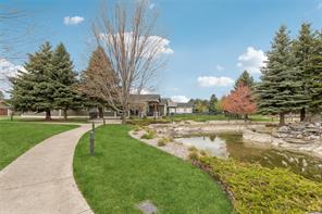 A curved sidewalk runs through green grass next to a small pond with rocks and shrubs—perfect for the HOT Summer Listing Season, with evergreen and leafless trees, nearby houses, and a blue sky completing the scene.