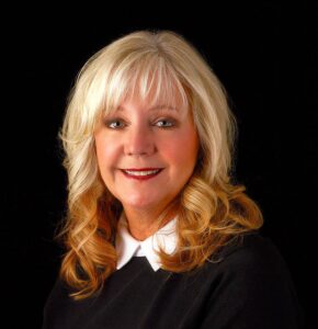 A woman with long blonde hair and bangs is smiling at the camera, wishing you a Merry Christmas. She is wearing a black top with a white collar against a solid black background.