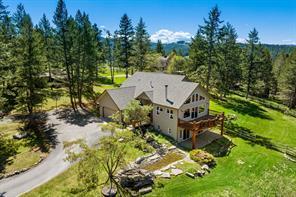 A large beige house with a wooden pergola sits among tall pines and green lawns, its curved driveway and stone landscaping inviting for the summer listing season, all under a bright blue sky.