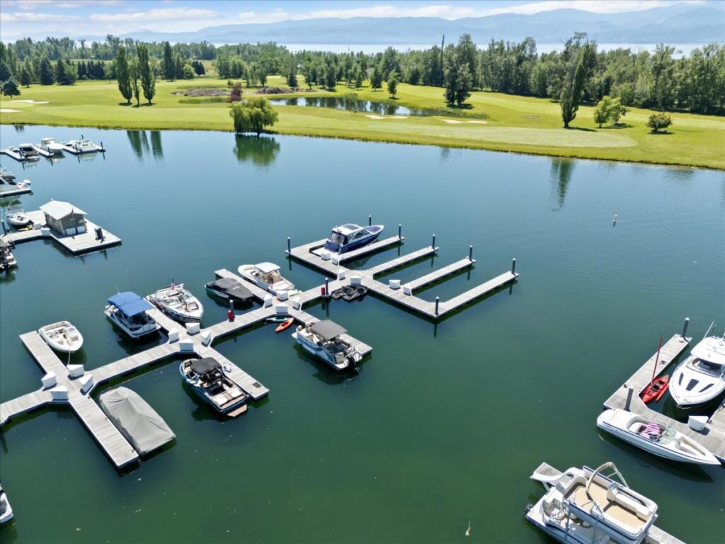 Aerial view of a marina with several boats docked at floating piers on calm water, perfect for Lake Days and July Journeys, surrounded by green grass, trees, and hills under a partly cloudy sky.