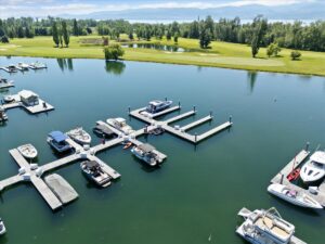 Aerial view of a marina with several boats docked at piers on calm water, surrounded by green grass and trees. September in Bigfork brings a golf course in the background under a partly cloudy sky as summer softens into fall.