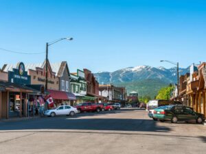 Small town main street lined with shops and parked cars on both sides; community lights glow softly as mountains with scattered snow patches rise in the background under a clear blue sky.