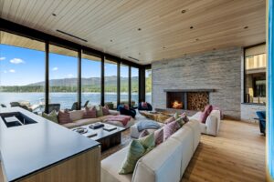 Spacious modern living room with large windows overlooking Flathead Lake and mountains, featuring light wood floors, a stone fireplace, and two cream sofas with colorful pillows. Discover refined comfort in this stunning Flathead Lake Home.