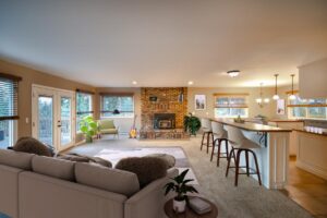 Spacious living area with a beige sectional sofa, brick fireplace, and green armchair radiates a cozy winter glow. Kitchen features a long bar and stools. Large windows fill the space with light. Plants, a guitar, and framed art complete this December real estate gem.