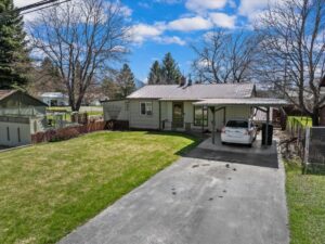 Single-story house with a front porch and attached carport, where a white car rests in the driveway. The yard, lush with green grass and mature trees, is perfect for June joys and new beginnings under a sky of scattered clouds.