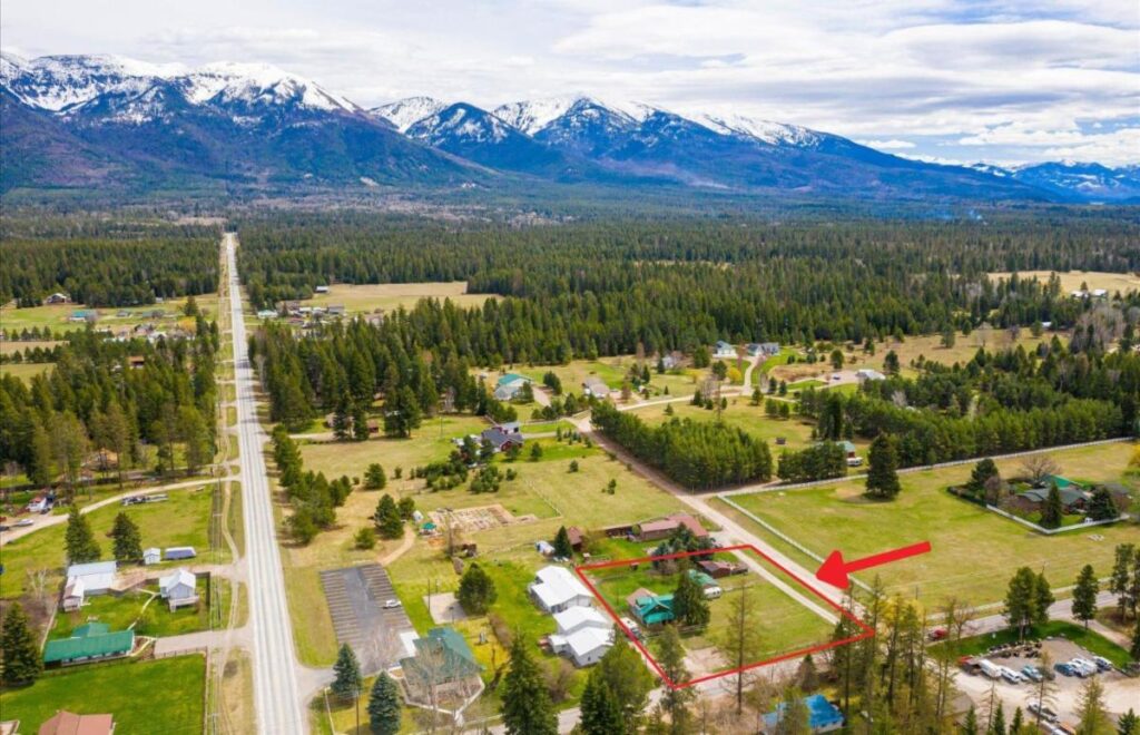 Aerial view of a rural neighborhood with scattered houses, green trees, and rolling open fields. Snow-capped mountains are in the background, and a red arrow points to a house within a fenced property in the foreground.